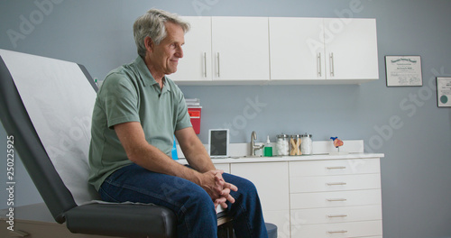 Senior Caucasian male patient waiting patiently for doctor while sitting on exam room table. Older man going to regular appointment for annual check up