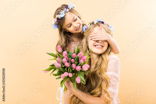 happy mother holding bouquet of pink tulips while cheerful child closing her eyes with hand on yellow background