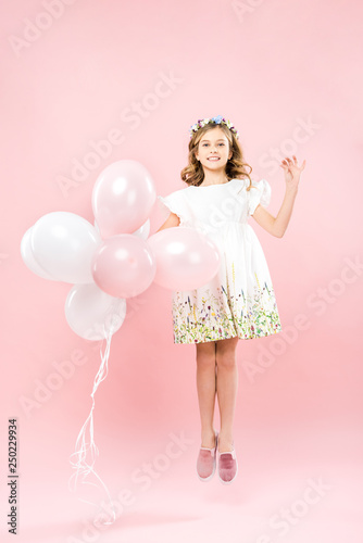 happy child in elegant white dress jumping with air balloons on pink background
