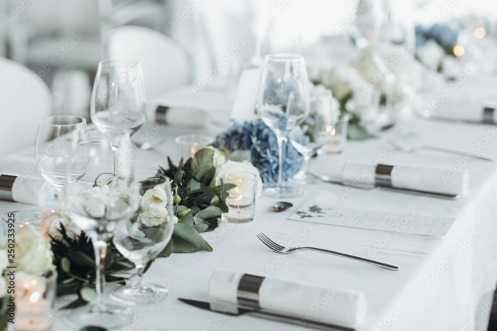 Wedding table decoration. Floral garland of greenery and blue flowers lies  between glasses on the white table Stock Photo | Adobe Stock