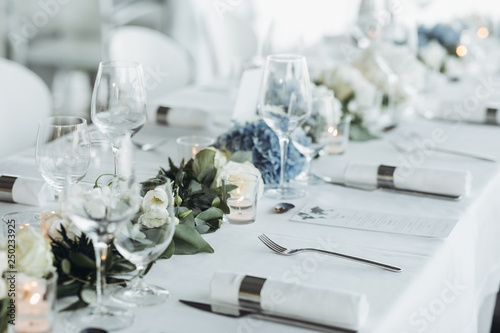 Wedding table decoration. Floral garland of greenery and blue flowers lies between glasses on the white table photo