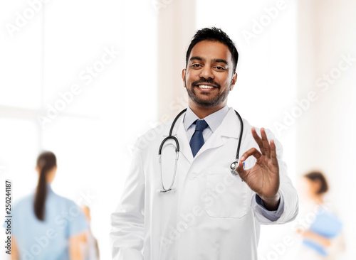 medicine, gesture and healthcare concept - smiling indian male doctor in white coat with stethoscope showing ok hand sign over hospital background