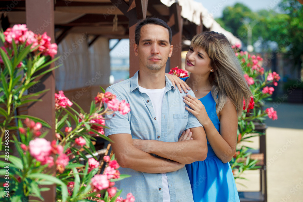 Obraz premium couple, guy and girl stand on a background of flowers, hug, happy. Guy looks at the camera, the girl looks at the guy, put her hands on his shoulders. On the street. Love story