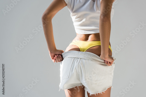 Schilderij op canvas Cropped view of woman in yellow panties taking off shorts on grey background