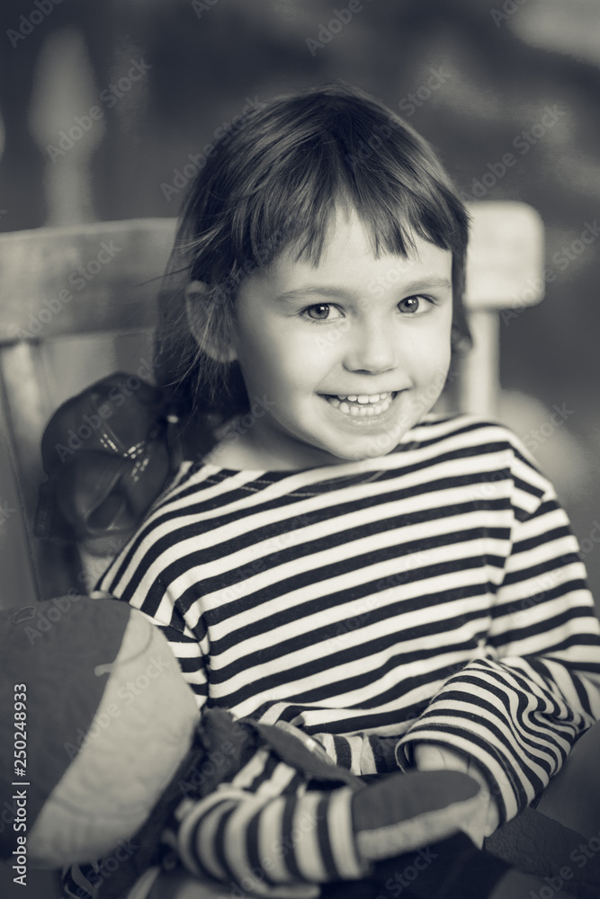A little happy smiling girl in a skirt and striped t-shirt sitting on a chair and holding an old toy, black and white A little happy smiling girl in a skirt and striped t-shirt sitting on a chair and holding an old toy, black and white