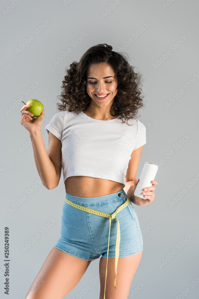 Laughing sexy girl with measuring tape holding green apple and beverage isolated on grey Laughing sexy girl with measuring tape holding green apple and beverage isolated on grey