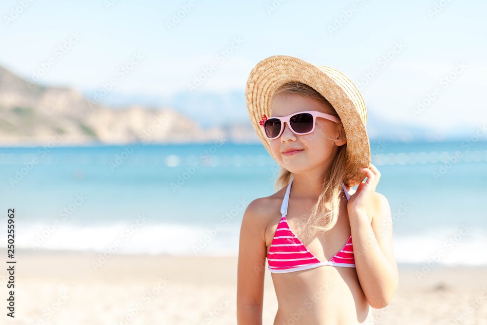 Beach girl in summer vacation. Kid at sea coast. Holiday, travel and summertime concept. Child in straw hat and sunglasses is happy tourist in resort outdoors. Cute lady is smiling, enjoying party.