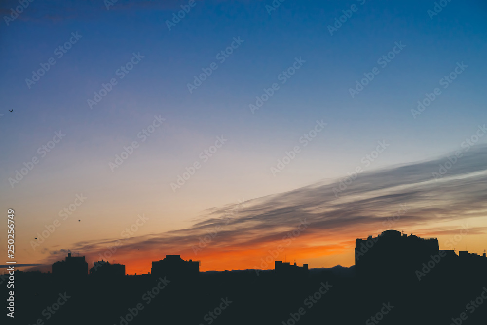 Cityscape with wonderful varicolored vivid dawn. Amazing dramatic blue cloud sky above dark silhouettes of city building roofs. Atmospheric background of orange sunrise in overcast weather. Copy space