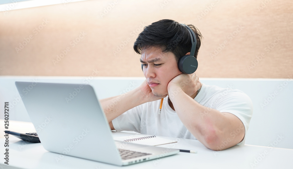 close up young asian student man wearing headset and looking at laptop display for learning mathematics online course from education website , writing lecture on notebook and solve lesson at library