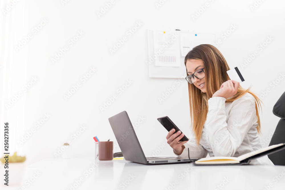 Pretty cheerful young manager woman buying with a bank card on mobile phone while sitting in her office. Pretty cheerful young manager woman buying with a bank card on mobile phone while sitting in her office.