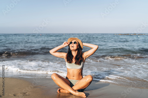 Pretty pan asian girl sitting on the beach near sea in hat and sunglasses.