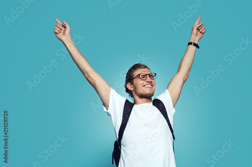 Portrait of a satisfied hipster boy with sunglasses and bagpack celebrating success, isolated over blue background. photo