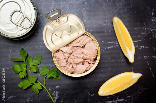 Opened tuna can with parsley and lemon slices on a black cutting board.