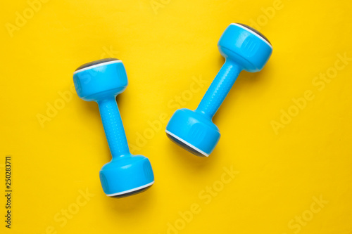 Two blue plastic dumbbells on yellow background Sport, fitness concept. Top view. Minimalism