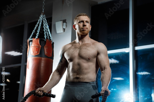 Strong man posing in gym with rope in his hands near the punching bag. photo