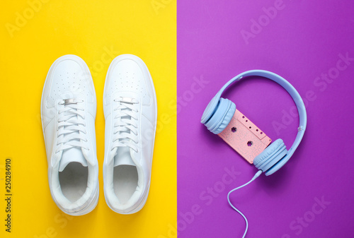 Retro style. 80s. Pop culture. Minimalismalism. Headphones with audio cassette, white sneakers against purple-yellow background. Top view