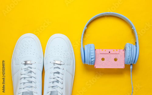 Retro style. 80s. Pop culture. Minimalismalism. Headphones with audio cassette, white sneakers on yellow background. Top view