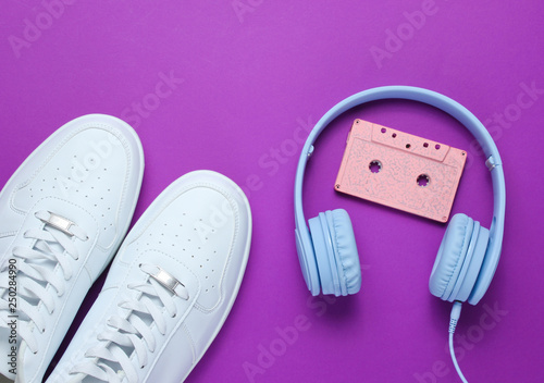 Retro style. 80s. Pop culture. Minimalismalism. Headphones with audio cassette, white sneakers on purple background. Top view