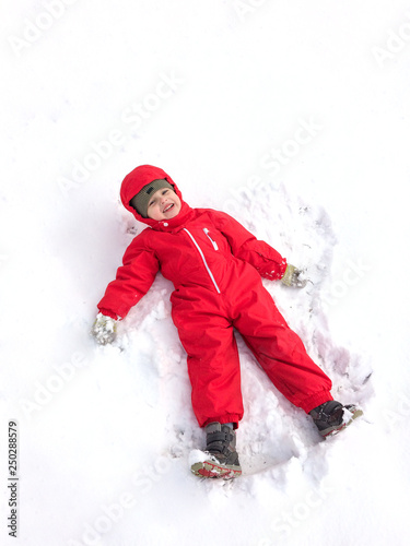 Cute little boy in a red winter jumpsuit in the winter on the street makes a snow angel and have fun.