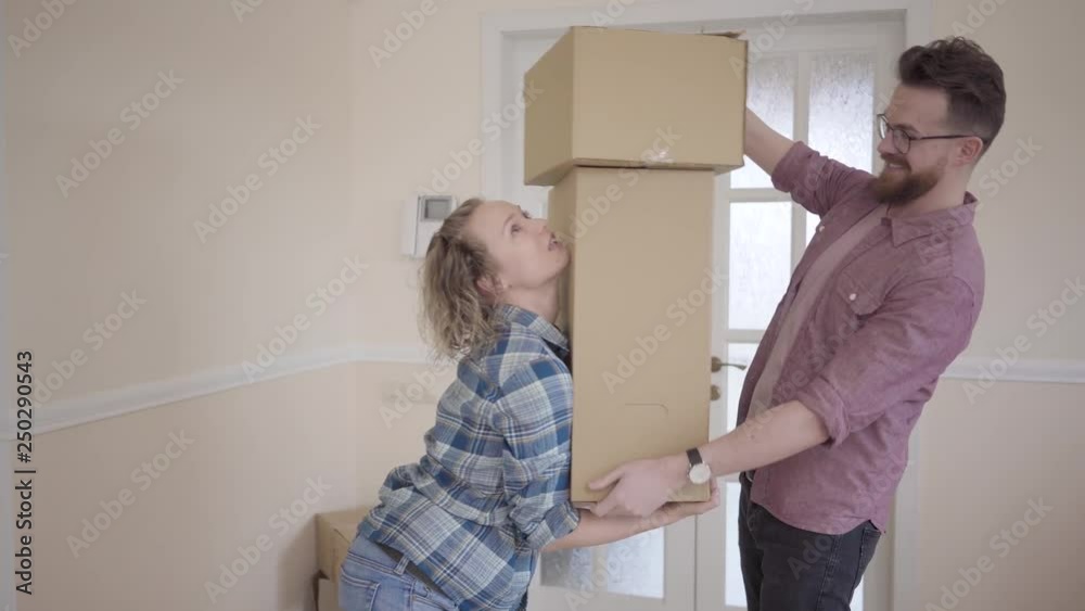 Tall man standing in the room with big box in hands. Woman takes the boxes from her husband and balances them in their arms. Married couple moves into a new home Tall man standing in the room with big box in hands. Woman takes the boxes from her husband and balances them in their arms. Married couple moves into a new home