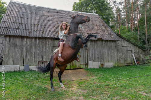 Girl astride horse, who costs on hinder legs, on background of old wooden barn. Riding lessons