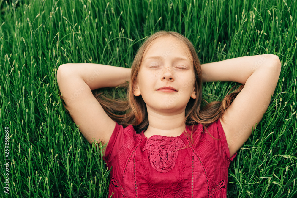 Cute little girl lying on bright green grass, holding arms behind head, eyes closed Cute little girl lying on bright green grass, holding arms behind head, eyes closed