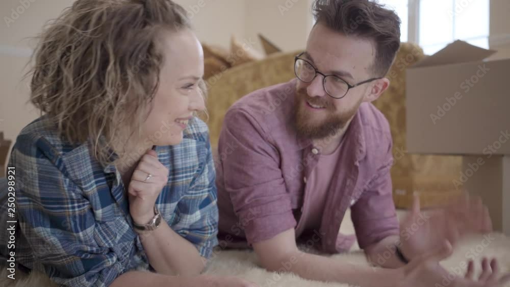 Young family lying on fluffy carpet close up with boxes in the background. Man showing key to woman, she stretches out hand and takes it, smiling. Married couple moves into a new home Young family lying on fluffy carpet close up with boxes in the background. Man showing key to woman, she stretches out hand and takes it, smiling. Married couple moves into a new home