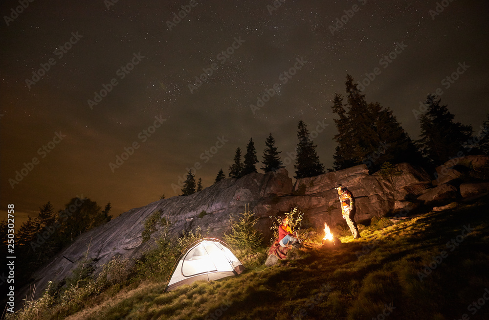 Night summer camping in the mountains. Happy couple travellers having a rest together beside campfire and illuminated tourist tent. On background big boulder, forest and beautiful night starry sky. - obrazy, fototapety, plakaty 