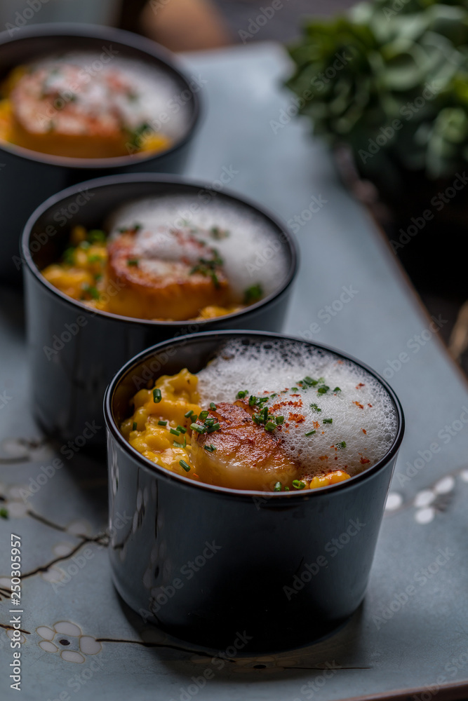 Orange pumpkin risotto with scallop and foamy sauce in black bowl on rustic wooden table