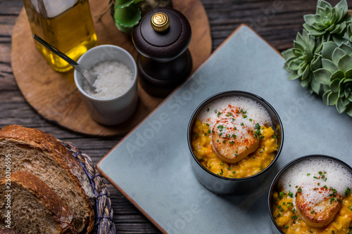 Orange pumpkin risotto with scallop and foamy sauce in black bowl on rustic wooden table