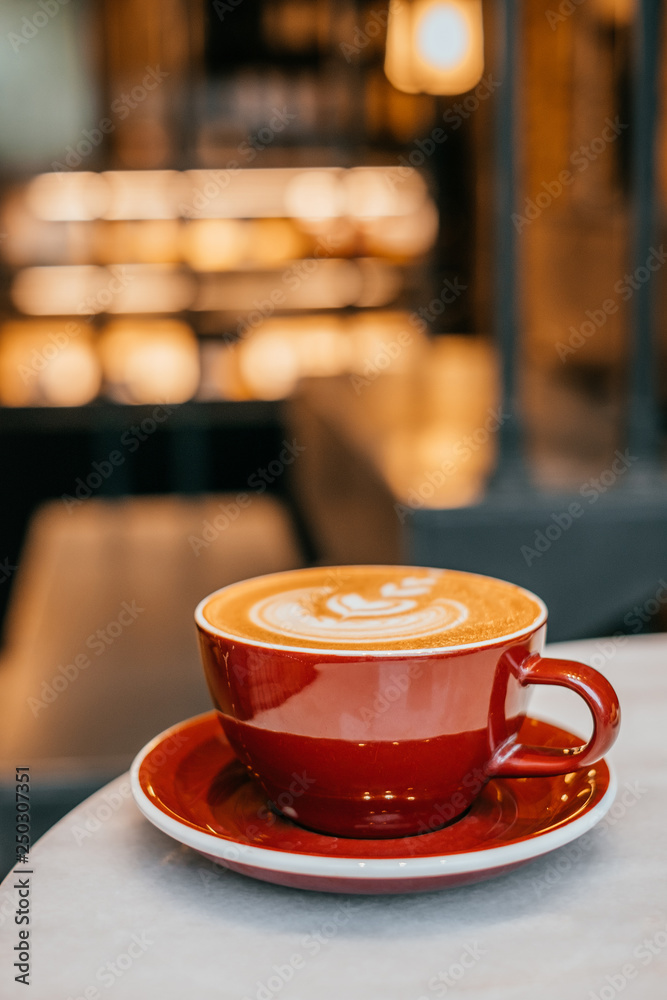 Red cup of cappuccino with beautiful latte art on round white marble table and blurred lights background. Red cup of cappuccino with beautiful latte art on round white marble table and blurred lights background.