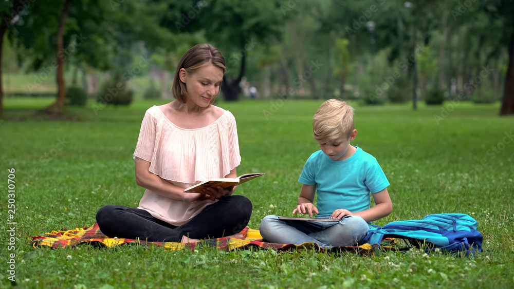 Mom reading book, son using tablet, alternative education with IT technologies Mom reading book, son using tablet, alternative education with IT technologies