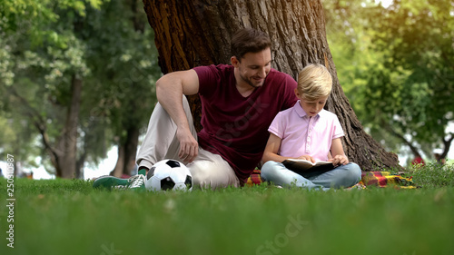 Tableau sur toile Dad and son reading book, sitting in park under tree, hobby, togetherness