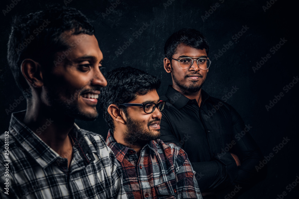 Close-up portrait of three happy Indian students wearing casual clothes against a dark textured wall.