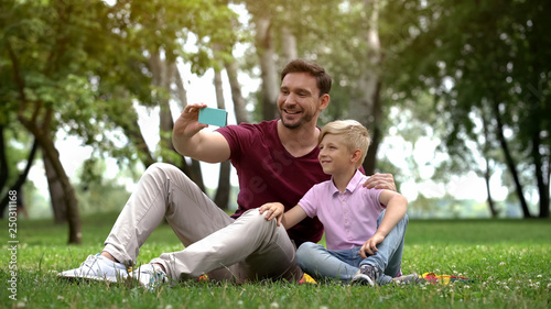 Dad and boy taking selfie on smartphone, resting in park at weekend, free time © motortion