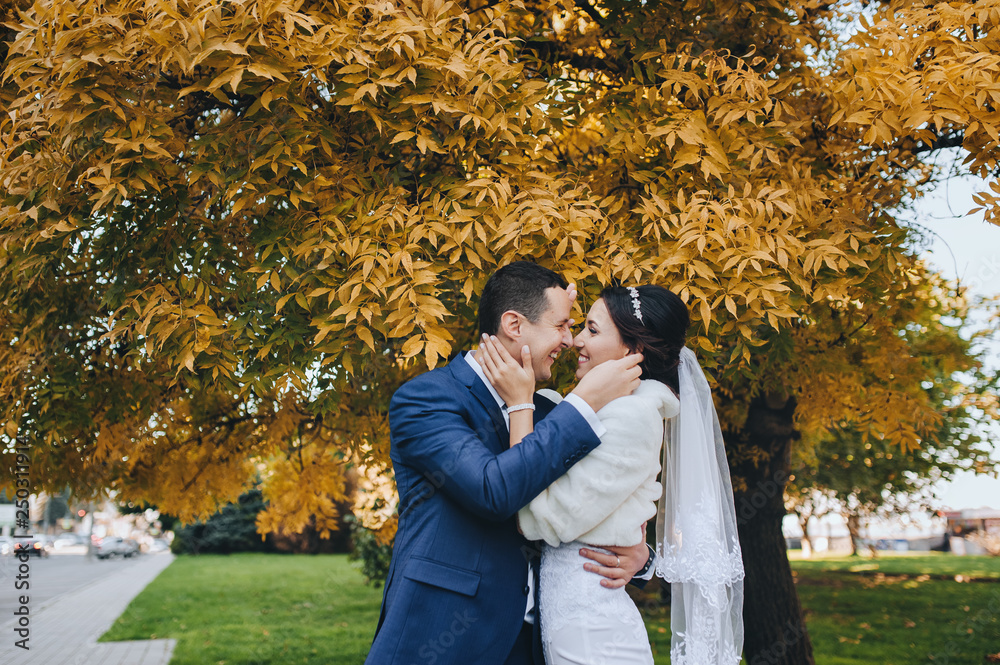 Beautiful and smiling newlyweds are hugging in the autumn in a park with yellow leaves. Portrait of a stylish groom with glasses and a cute bride in a white dress. Wedding photography.