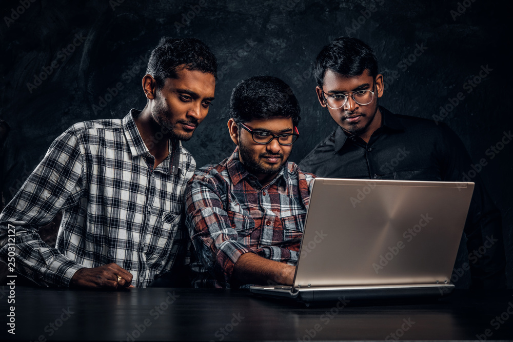 Three Indian students working on a project together standing at the table with a laptop in a dark room Three Indian students working on a project together standing at the table with a laptop in a dark room