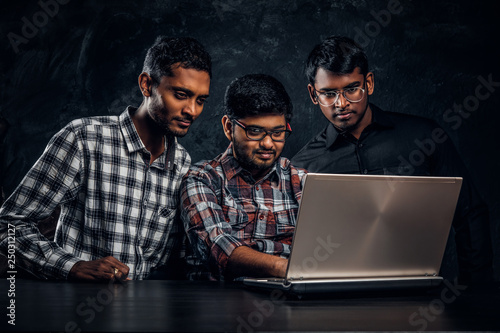 Three Indian students working on a project together standing at the table with a laptop in a dark room photo