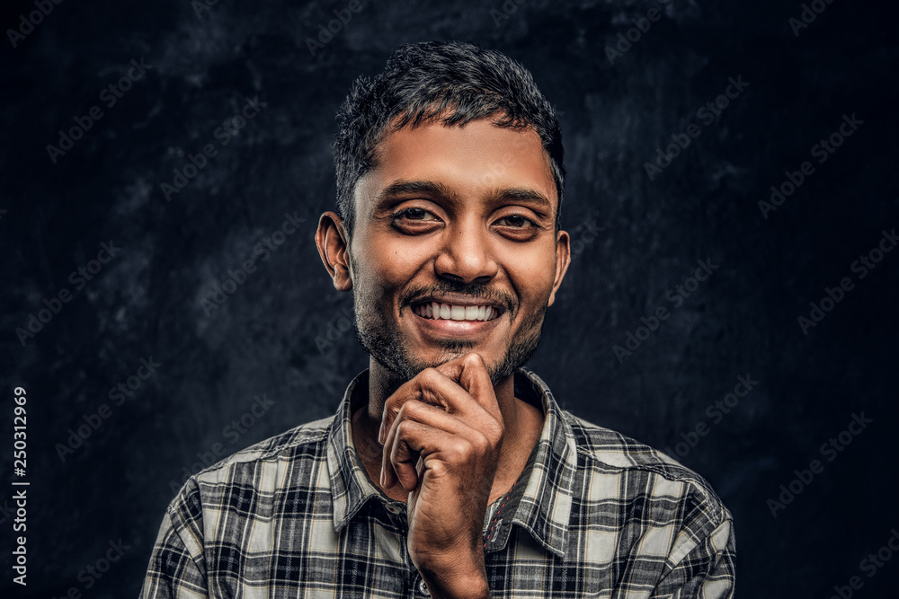 Fototapeta premium Portrait of a young Indian guy wearing a checkered shirt holding hand on chin, smiling and looking at a camera in a studio