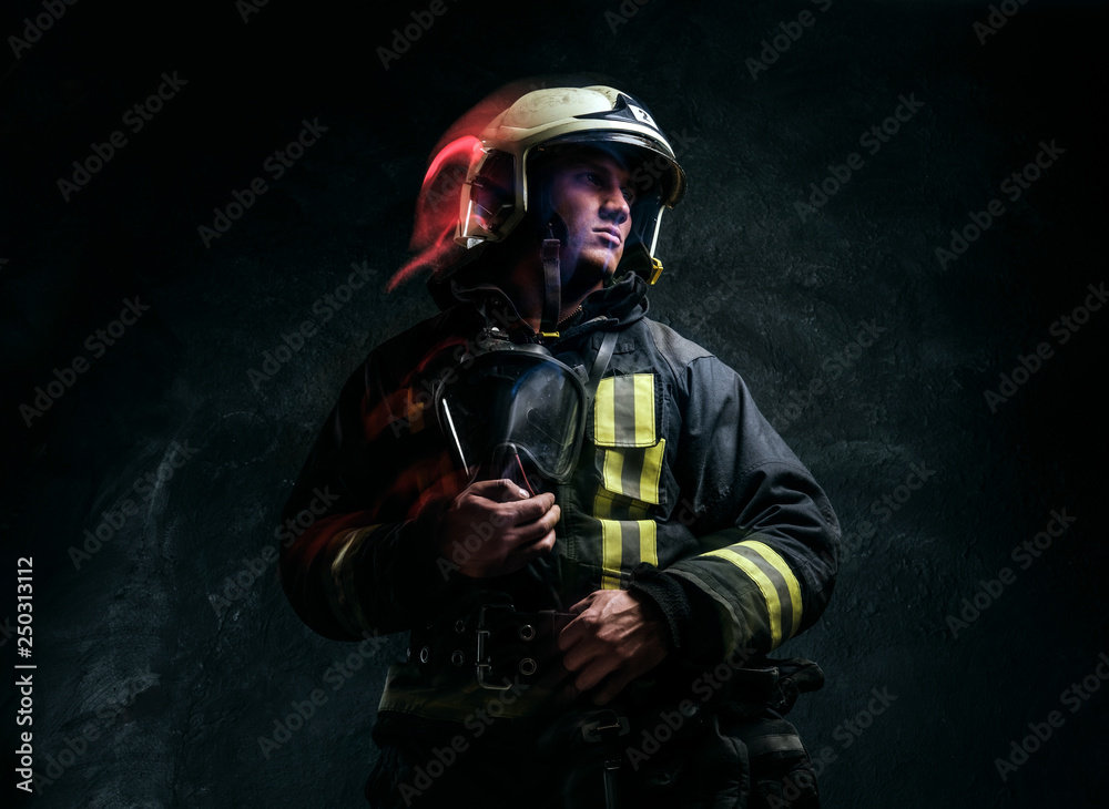 Studio portrait in a dark studio against a textured wall. Brutal firefighter in uniform and safety helmet holding an oxygen mask and looking sideways with a confident look.