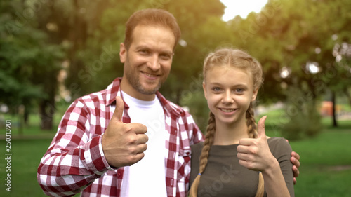 Cheerful father and teenage daughter smiling into camera, showing thumbs up