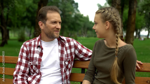 Beautiful teenage daughter telling secrets to father, relaxing on bench in park © motortion