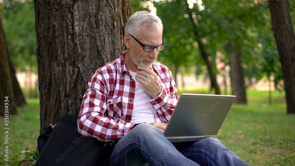 Elderly man sitting on grass and chatting online on laptop, dating applications Elderly man sitting on grass and chatting online on laptop, dating applications