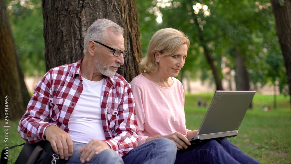Male and female pensioners choosing hotel for vacation on laptop resting in park Male and female pensioners choosing hotel for vacation on laptop resting in park