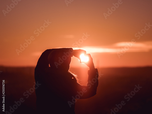 Image of woman from behind forming a heart with her hands with sun shining towards her face