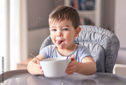 Obraz na plátně Adorable thankful little baby boy with smeared face and tongue out just finished tasty meal and hold white bowl looking at camera wanting more sitting at high feeding chair