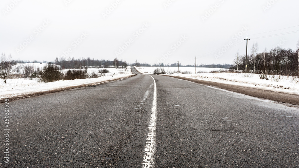 Rural asphalt road in winter. Dirty road with cracks. Winter fields and forest on a cloudy day