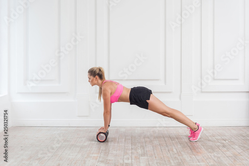 Profile side view portrait of athletic blonde young woman in fit wear using foam roller in gym to workout to loss weight and doing plank. Rehabilitation sports medicine. Indoor, studio shot,