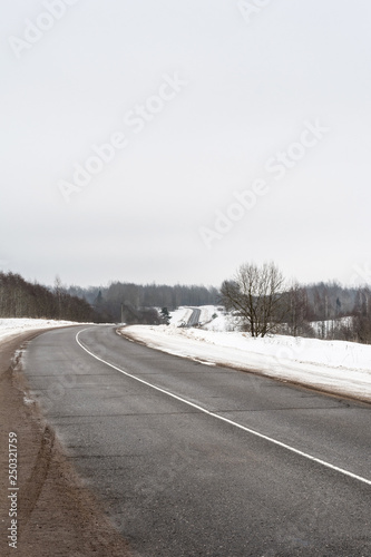 Rural asphalt road in winter. Dirty road with cracks. Winter fields and forest on a cloudy day