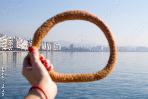 womans hand with red maniqure holding the greek bagel koulouri with sea and city on the background